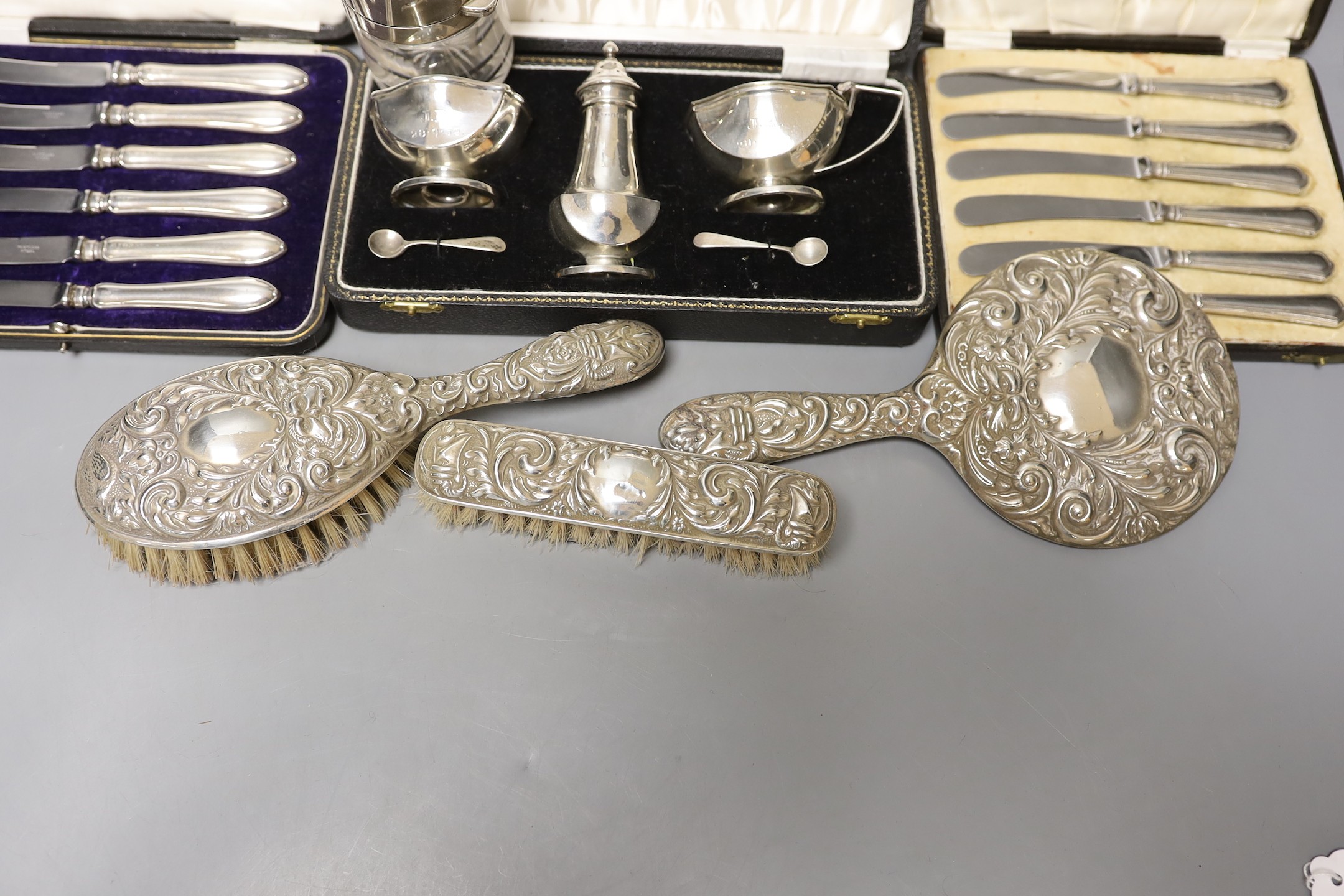 A cased 1950's silver three piece condiment set, two cased sets of six silver handled tea knives, a silver mounted glass salts bottle, silver mounted hand mirror and two brushes.